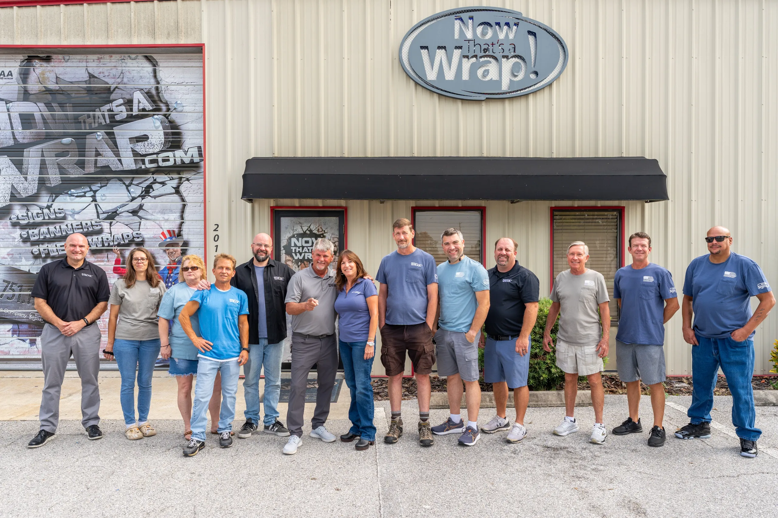 A group of 13 people stand smiling in front of a building with a sign reading “Now that’s a Wrap!” on the exterior wall.