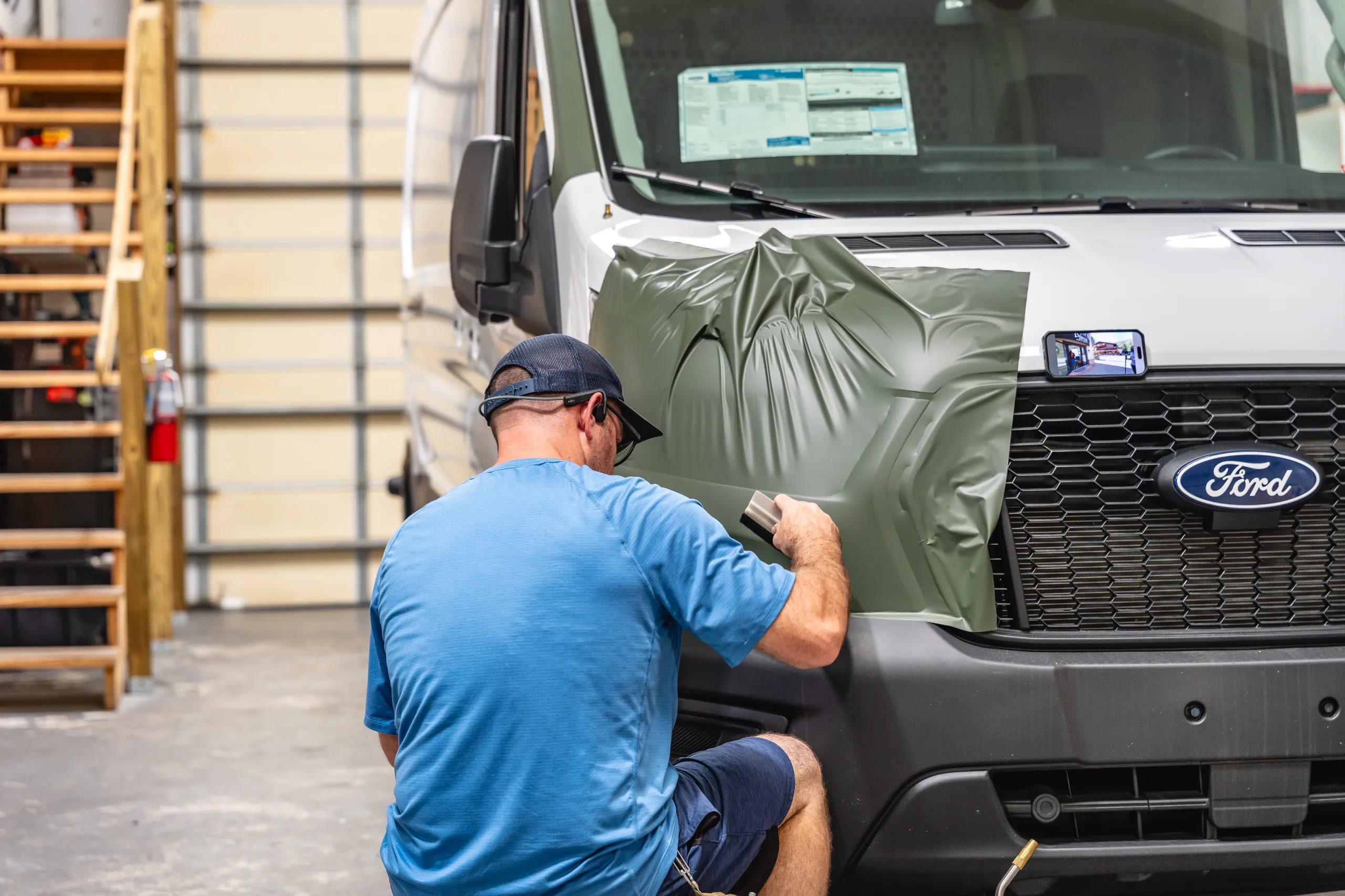 Man in blue shirt applies a green vinyl wrap to the hood of a Ford van inside a workshop.