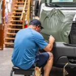 A person applies a green vinyl wrap to the front of a Ford van inside a workshop.
