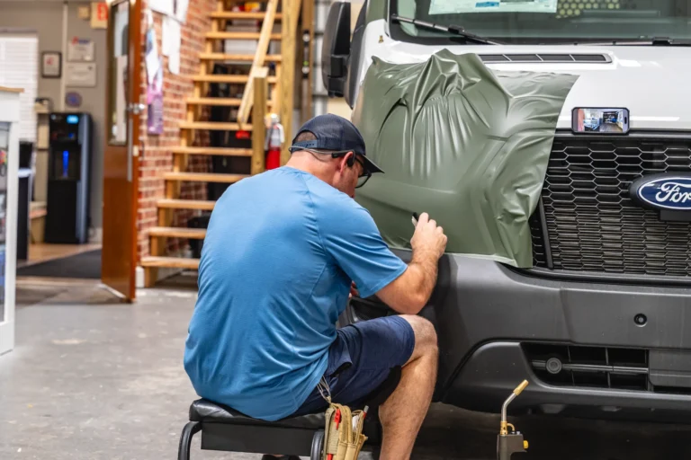 A person applies a green vinyl wrap to the front of a Ford van inside a workshop.