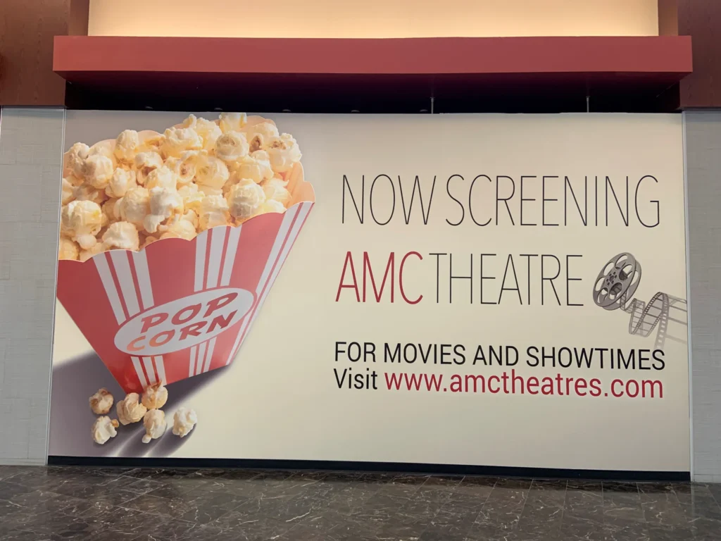 Large sign showing a popcorn tub and film reel, reading “Now screening AMC Theatre. For movies and showtimes visit www.amctheatres.com.”.