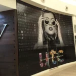 Large wall mural of a woman holding sunglasses, with brand names in the background and the words “BE THE CENTER OF ATTENTION” below colorful “UTC” letters at a shopping mall.