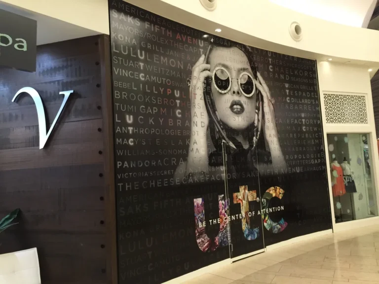 Large wall mural of a woman holding sunglasses, with brand names in the background and the words “BE THE CENTER OF ATTENTION” below colorful “UTC” letters at a shopping mall.