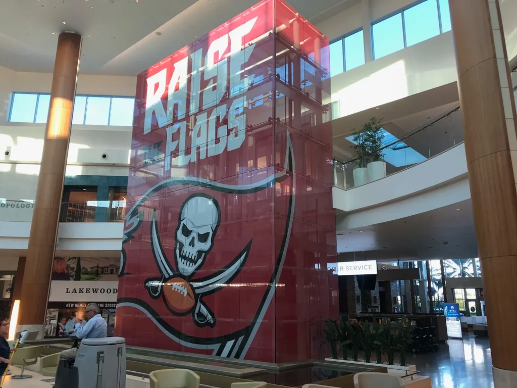 A tall glass display in a mall features a large Tampa Bay Buccaneers logo and the words “RAISE FLAGS” in bold white letters.