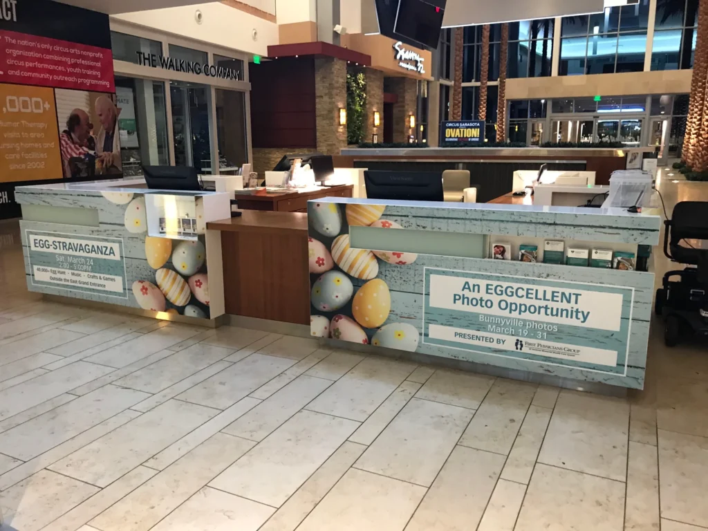 Shopping mall information desk decorated with pastel Easter egg designs, signs for an egg-themed photo opportunity, and event details displayed on the front panels.