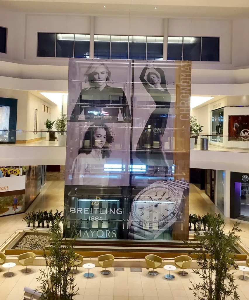 Large mall atrium with a tall vertical display featuring photos of models and a luxury watch advertisement; empty yellow chairs and tables are arranged below.