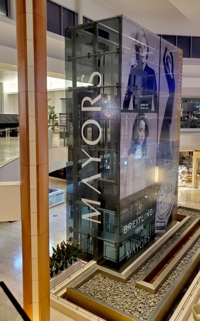 Glass elevator shaft in a shopping mall with “MAYORS” branding and luxury watch advertisements, surrounded by pebble landscaping and retail stores in the background.