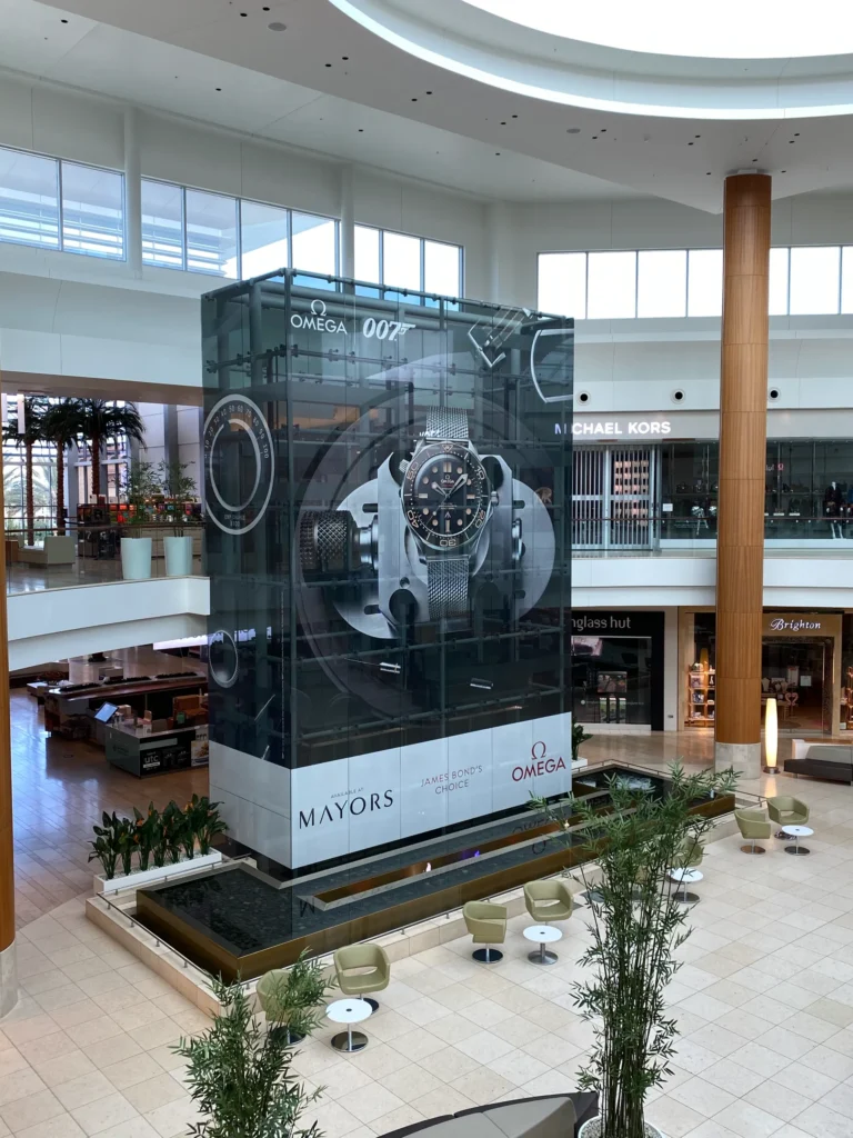 Large Omega watch advertisement display stands in the center of a shopping mall, surrounded by stores, seating, plants, and natural light from skylights above.