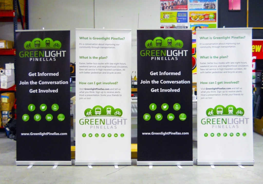 Four vertical Greenlight Pinellas banners display information about the project, ways to get involved, and social media icons, set up indoors with colorful posters in the background.