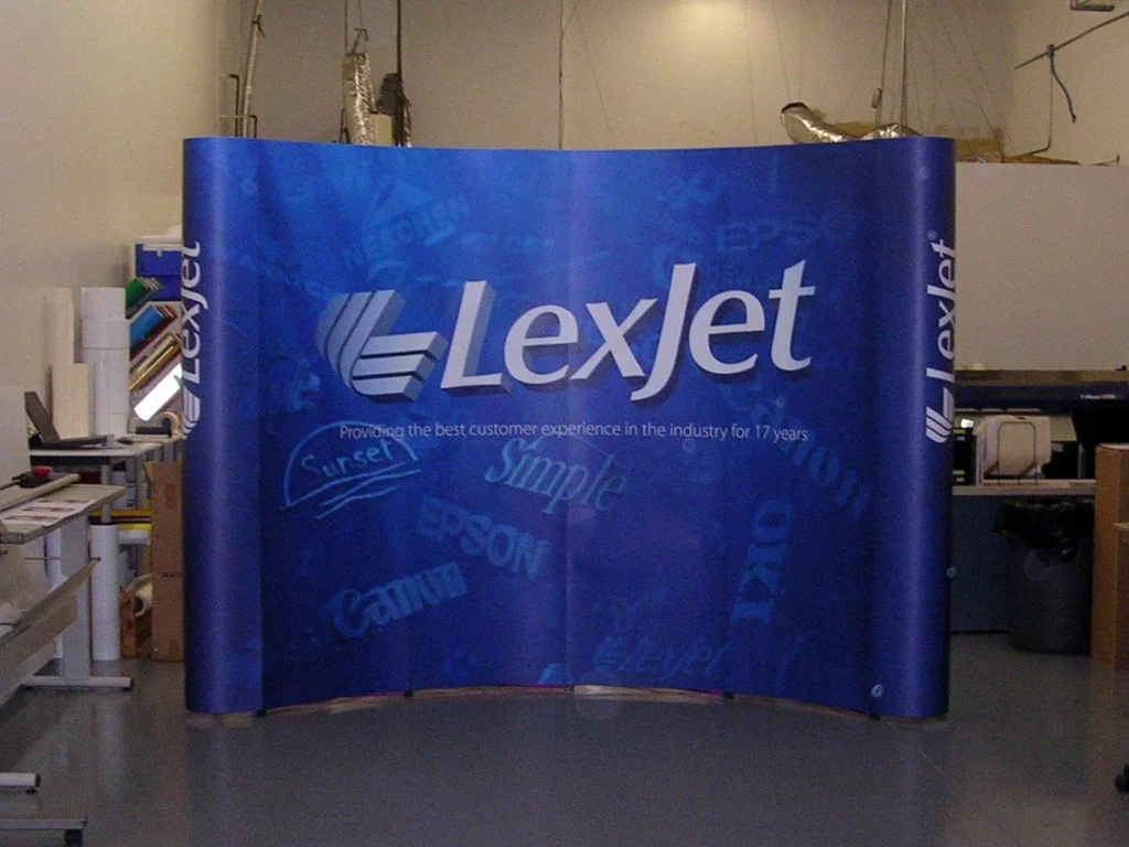 A blue LexJet trade show display stands in a workshop, featuring the LexJet logo and slogan, with various brand names faintly visible in the background.