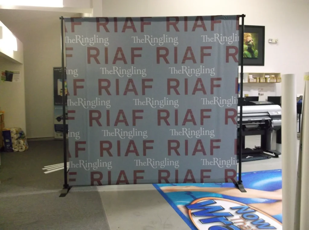 A printed backdrop displays “RIAF” and “The Ringling” repeatedly in red and white text, set up in a print shop with equipment and posters visible in the background.