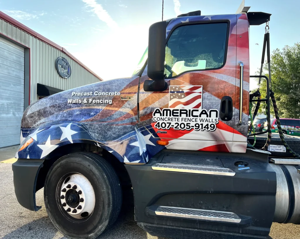 Semi-truck with an American flag design advertising “American Concrete Fence Walls” and phone number, parked outside a building.