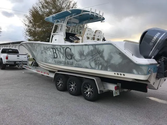 A white and gray boat with “BWC BLUEWATER GEAR” lettering is loaded on a triple-axle trailer, hitched to a pickup truck in a parking lot.