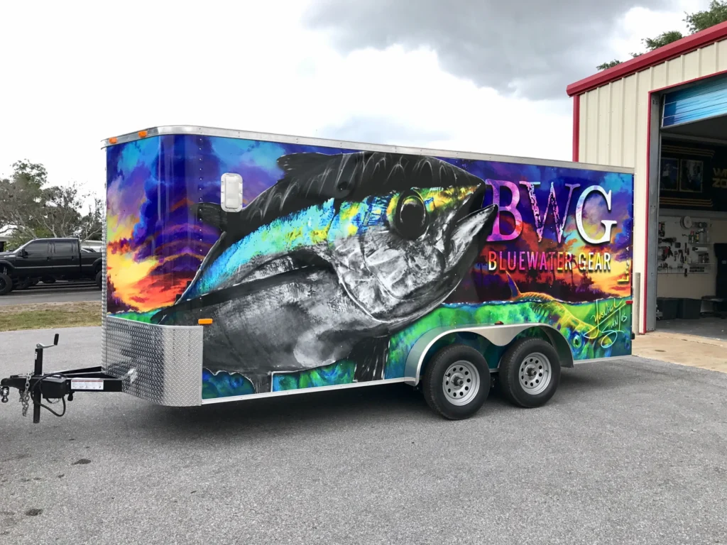 A trailer with a vivid mural of a large fish and the text “BWG Bluewater Gear,” parked outside a garage.