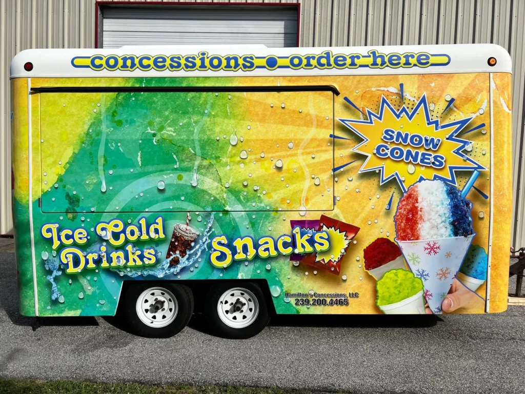 A colorful concession trailer advertises ice cold drinks, snacks, and snow cones with bright graphics and text, parked outside a building.