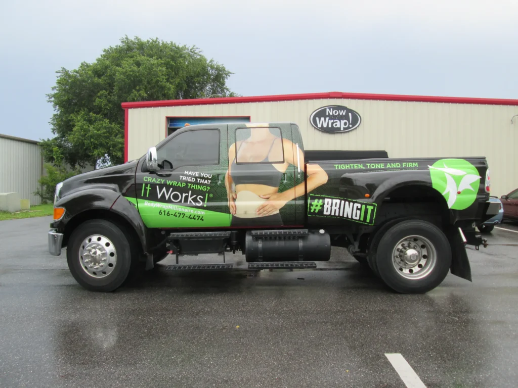 Large pickup truck parked outside a building, featuring an advertisement for a wrap product with the slogan “It Works!” and an image of a wrapped torso on the vehicle’s side.