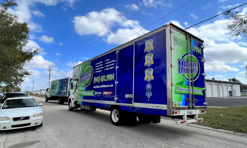 Two large moving trucks with bright blue and green graphics are parked on a street under a partly cloudy sky; a white car is parked nearby.