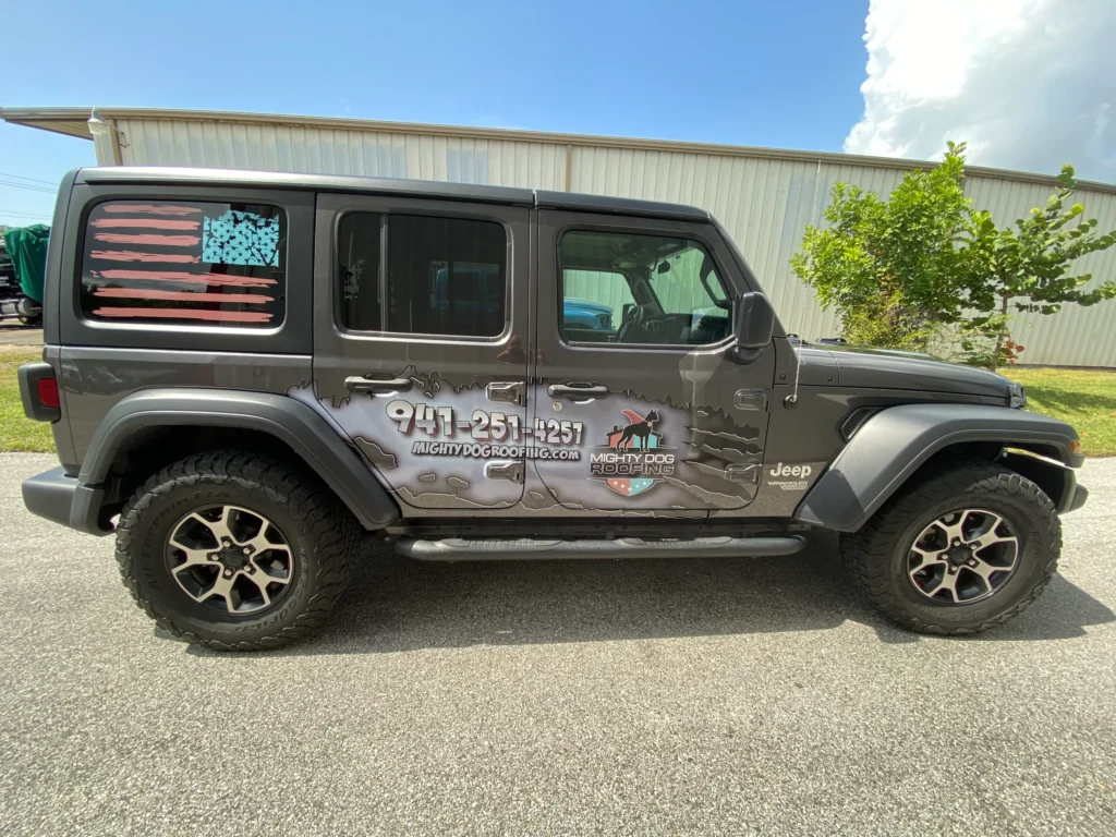 Gray Jeep with custom decals, including a stylized American flag on the rear window and a business logo with contact information on the door, parked on pavement near a metal building.
