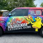 A colorful van with “Mellow Mushroom Catering Cube” branding, cartoon mushroom, menu items listed, a QR code, and contact number parked in a lot.