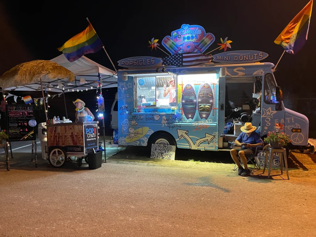 A brightly lit ice cream and mini donut food truck at night, decorated with rainbow flags, with two people sitting and one person serving from a small cart.
