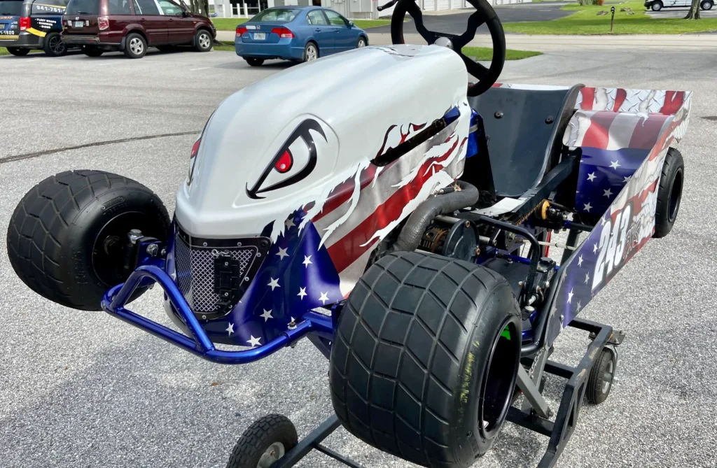 Custom go-kart with large tires, American flag design, and a painted fierce face on the front, parked on a trolley in a lot.