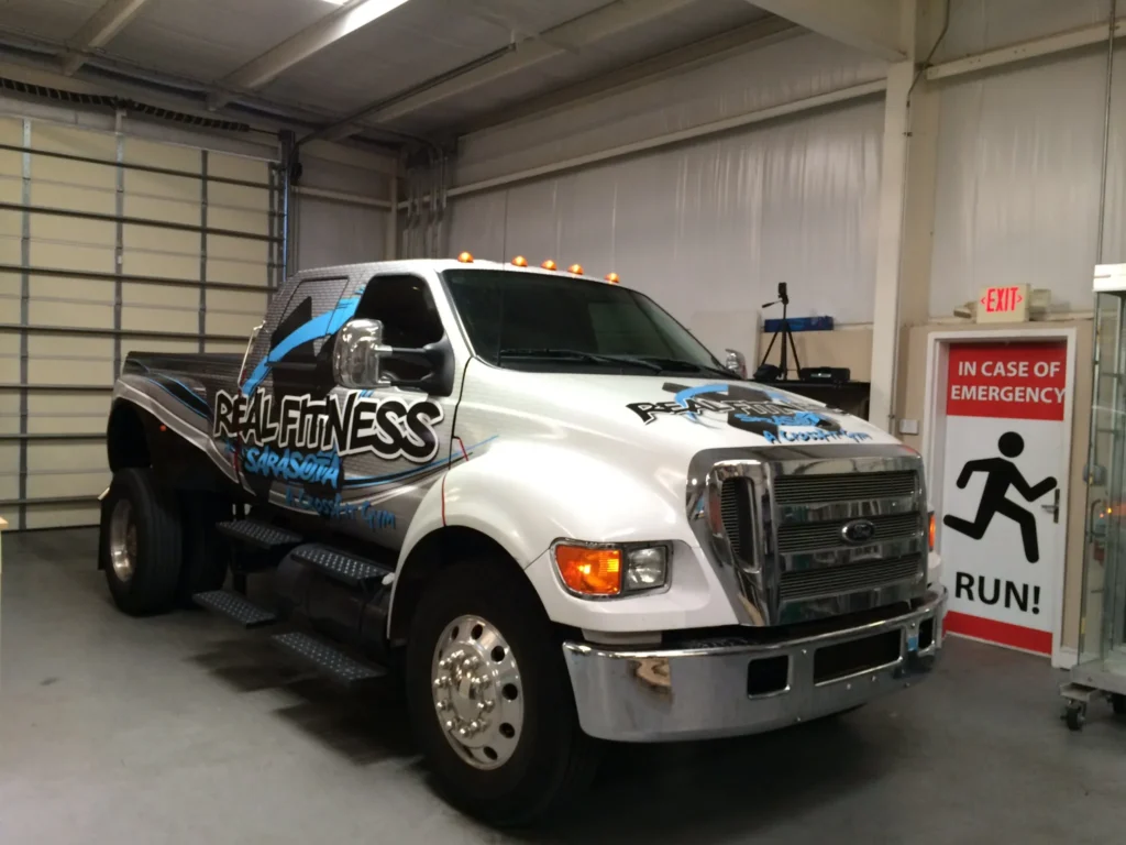 A large white and blue truck with “Real Fitness Sarasota” branding is parked inside a garage near an emergency exit sign that says “RUN!”.