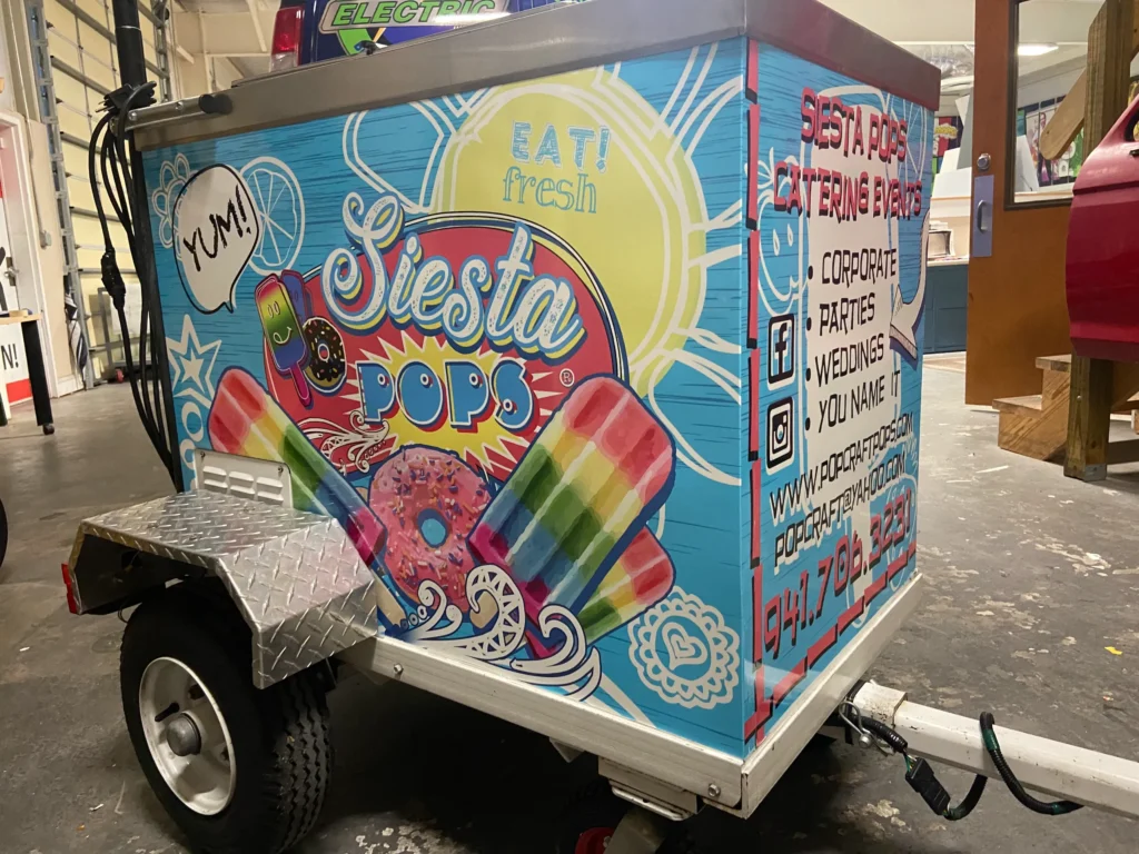 A colorful Siesta Pops cart with illustrations of rainbow popsicles and donuts, advertising catering for events, with contact info and social media details on the side.