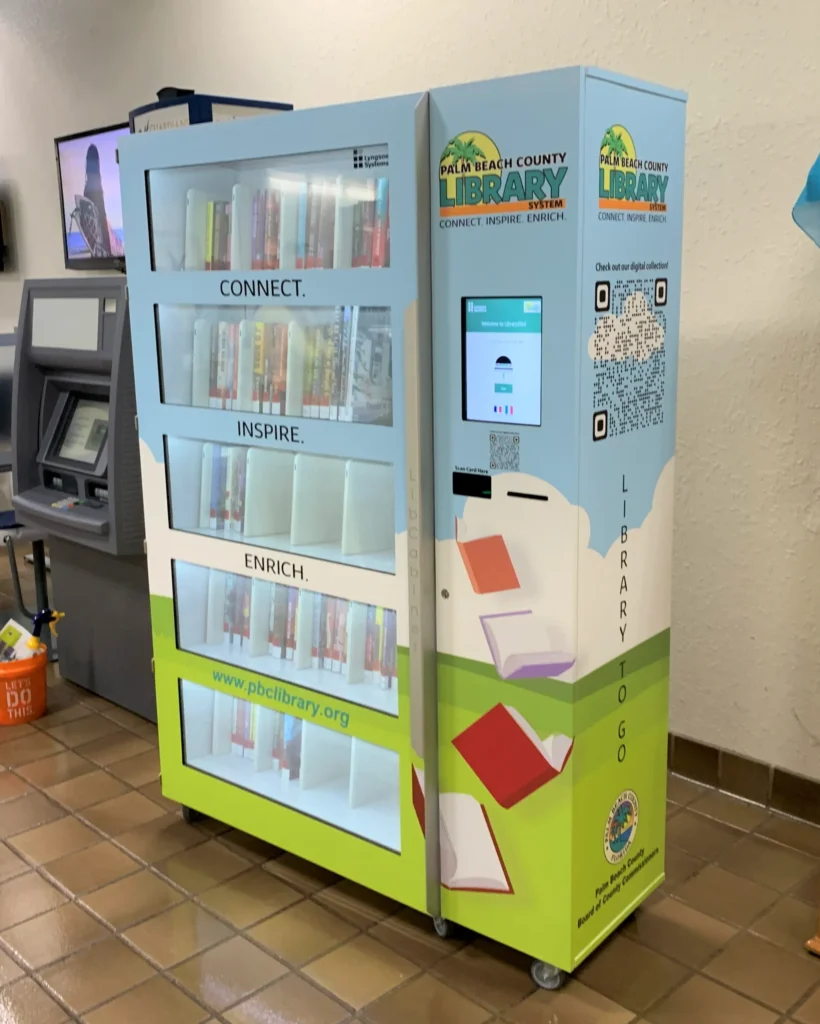 A Palm Beach County Library vending machine filled with books, featuring a touchscreen for borrowing and returning, next to an ATM on a tiled floor.