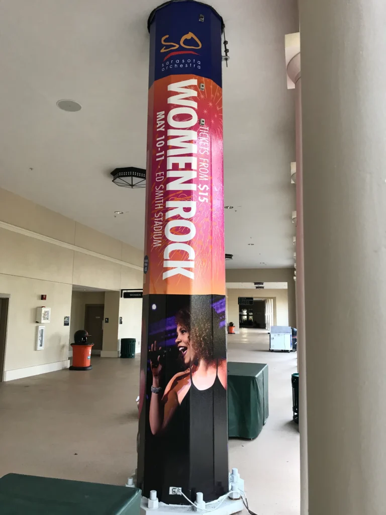 A tall column with a “Women Rock” event poster featuring a singer with a microphone, event date May 10–11, at Ed Smith Stadium, and ticket prices starting at $15.