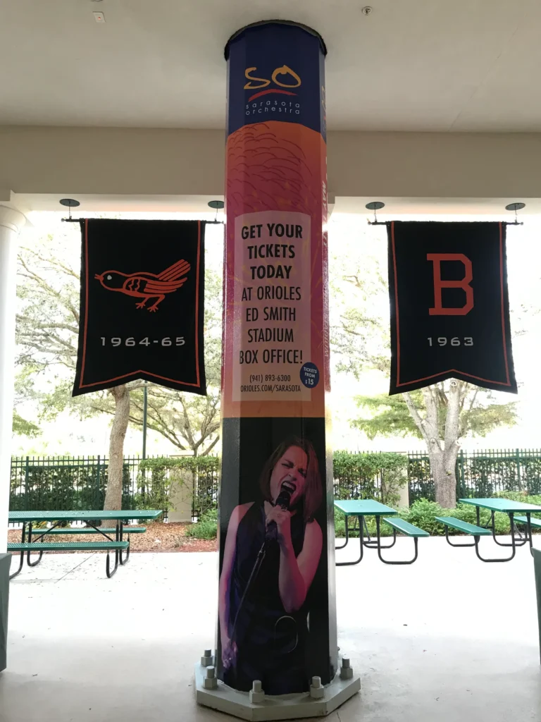 A stadium pillar displays a concert ad featuring a female singer, flanked by Orioles and “B” team banners with years 1964-65 and 1963. Benches and trees are visible in the background.