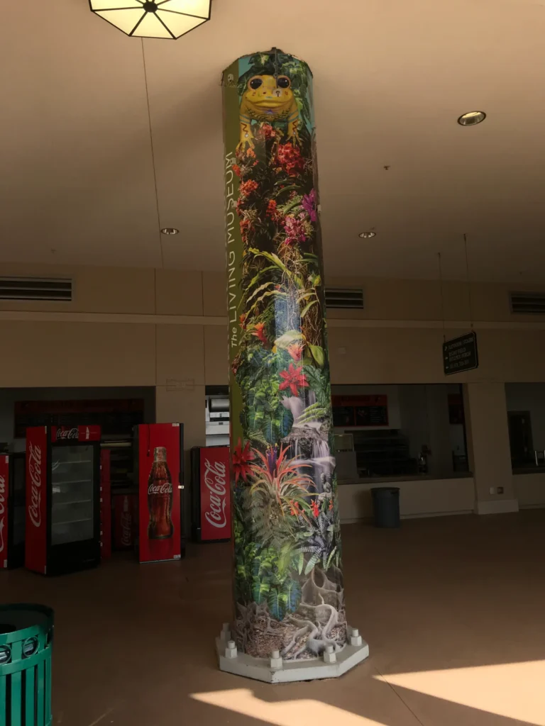 A cylindrical column is wrapped in a colorful rainforest mural with plants, vines, and a frog at the top. Coca-Cola vending machines are visible in the background.