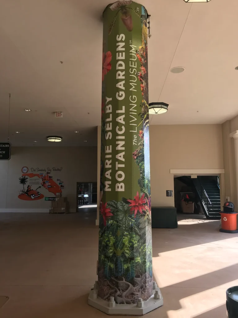 A pillar wrapped in a Marie Selby Botanical Gardens advertisement stands inside a building lobby with stairs and signage in the background.