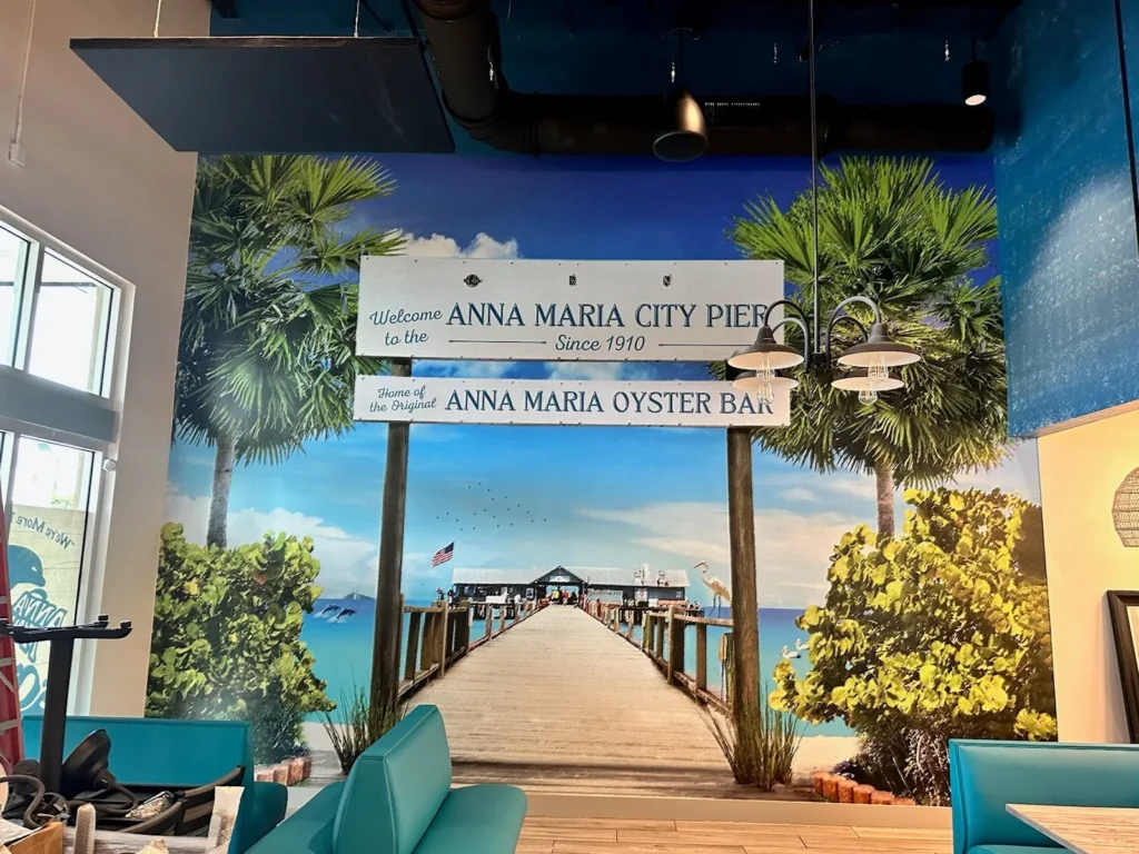 Wall mural of Anna Maria City Pier with palm trees, a wooden pier over blue water, and a sign reading “Anna Maria City Pier” and “Anna Maria Oyster Bar.” Blue seating in foreground.