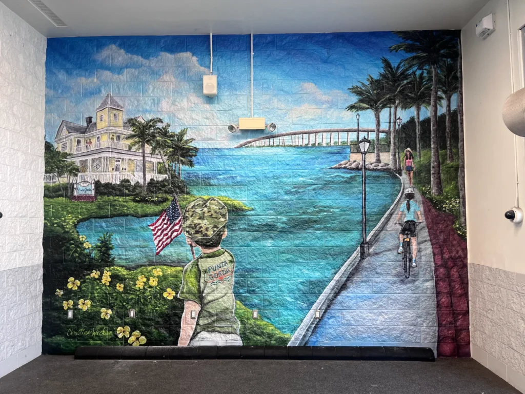 Mural of a child holding an American flag, facing a waterfront path with cyclists, palm trees, and a large house near a bridge under a blue sky.